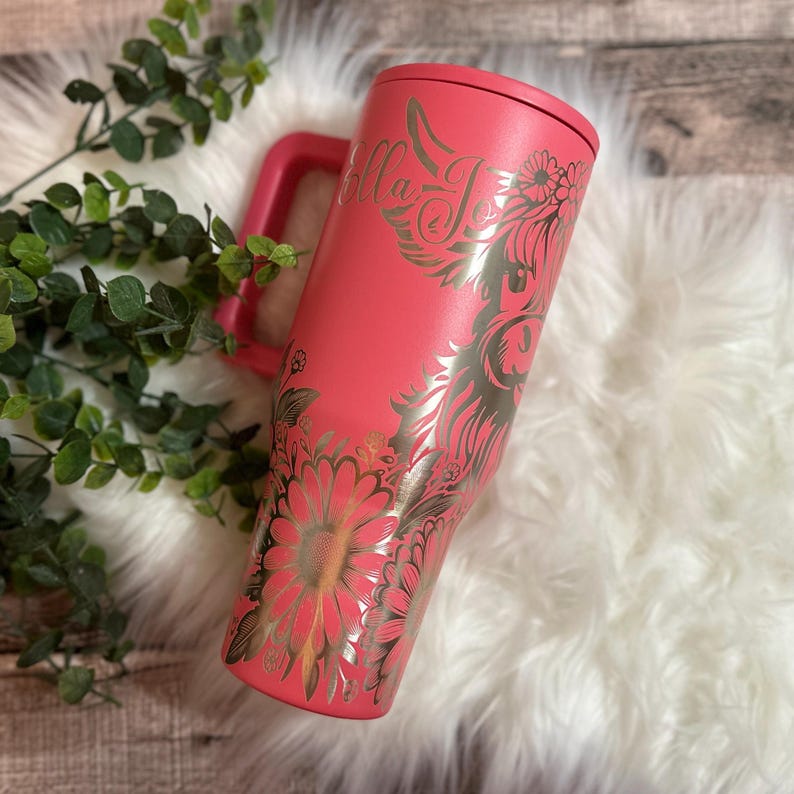 May include: A coral-colored tumbler with a handle and a floral design. The tumbler features the name "Bella Jo" in a decorative font. The design includes a cow silhouette and flowers in a metallic gold color. The tumbler is on a white fluffy surface.