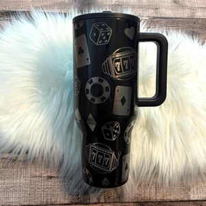 May include: Black travel mug with a handle, decorated with casino-themed graphics. The design includes dice, playing cards, poker chips, and a '777' jackpot symbol. The mug is cylindrical and appears to be made of stainless steel.