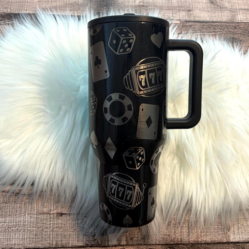 May include: Black travel mug with a handle, decorated with casino-themed graphics. The design includes dice, playing cards, poker chips, and a '777' jackpot symbol. The mug is cylindrical and appears to be made of stainless steel.