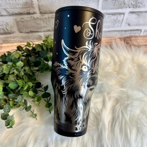 May include: Black stainless steel tumbler featuring a detailed silver engraving of a Highland cow. The tumbler has the name "Sha" in a cursive font, along with a heart and stars. The tumbler is designed to keep beverages cold or hot.