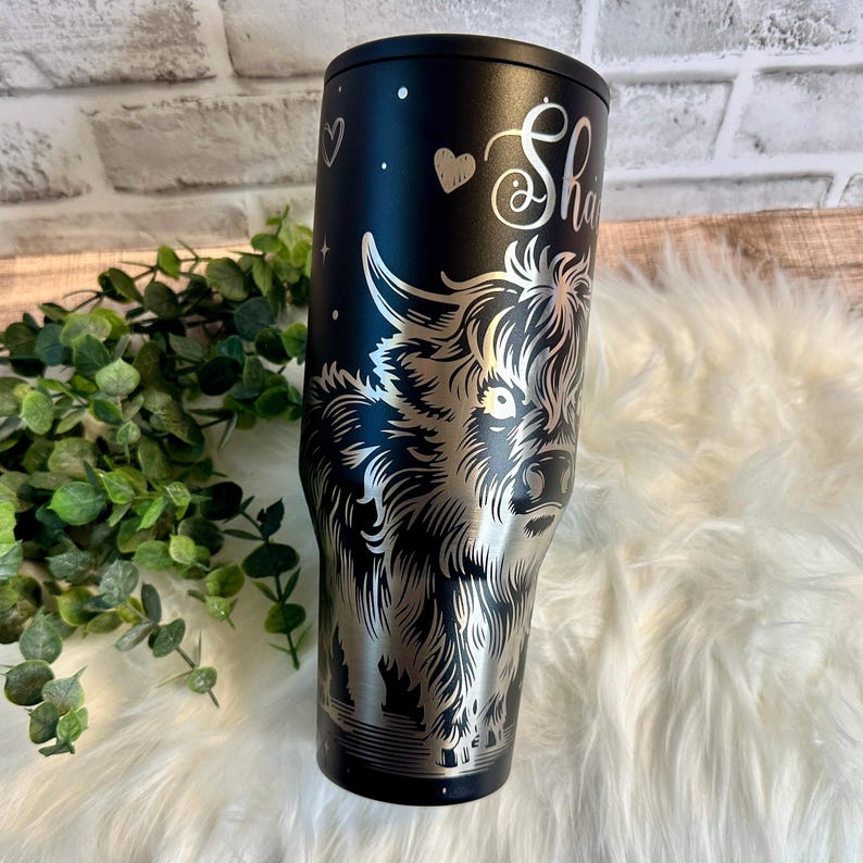 May include: Black stainless steel tumbler featuring a detailed silver engraving of a Highland cow. The tumbler has the name "Sha" in a cursive font, along with a heart and stars. The tumbler is designed to keep beverages cold or hot.