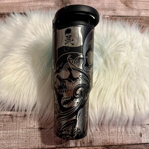 A black and silver tumbler mug with a skull and crossbones design, placed on a white furry surface.