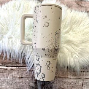 A white ceramic mug with a black and white illustration of a bear sitting in a pond, surrounded by bubbles, is placed on a white fur rug.