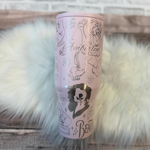 Puede incluir: Un vaso de color rosa claro con tapa plateada e ilustraciones plateadas de personajes y rosas. El vaso tiene el nombre "Amberlyn" y la palabra "Beauty" en escritura plateada. El vaso es cil&iacute;ndrico y parece ser de acero inoxidable.