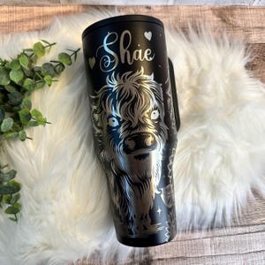 May include: Black tumbler with a silver engraved Highland cow design. The name "Shae" is written in cursive with heart accents. The tumbler has a handle and a black lid. The design is detailed and features stars and floral elements.