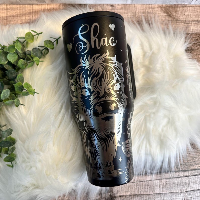 May include: Black tumbler with a silver engraved Highland cow design. The name "Shae" is written in cursive with heart accents. The tumbler has a handle and a black lid. The design is detailed and features stars and floral elements.