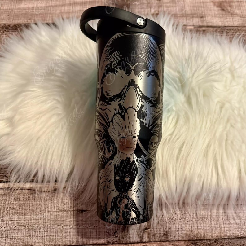 A black tumbler mug with a skull design and a handle, placed on a white furry surface.