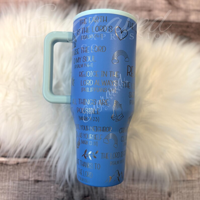 A blue tumbler mug with various Bible verse references and illustrations printed on it, placed on a white fur surface.
