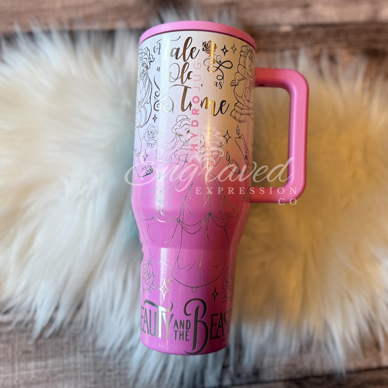 A pink and white tumbler mug with the text "Beauty and the Beast" printed on it, sitting on a white fur surface.
