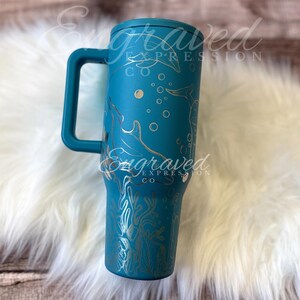 Puede incluir: Vaso t&eacute;rmico aislado de color turquesa con asa, con un dise&ntilde;o grabado de delfines, burbujas y corales. El vaso tiene una tapa turquesa a juego y las palabras "Engraved Expression Co" son visibles en el lateral.