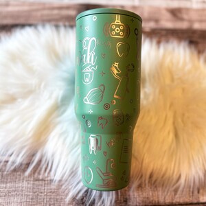 A green tumbler mug with various dental-related icons and text printed on it, placed on a white fur surface.