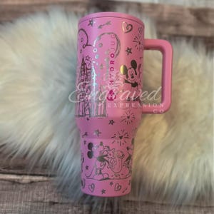 A pink tumbler mug with a Disney-themed design, featuring Mickey Mouse and other Disney characters, is placed on a white fur surface.