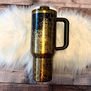 May include: A black and gold Stanley tumbler with a black handle. The tumbler features a bee and honeycomb design. The word "STANLEY" is printed vertically on the side of the tumbler. The tumbler is sitting on a white fluffy surface.