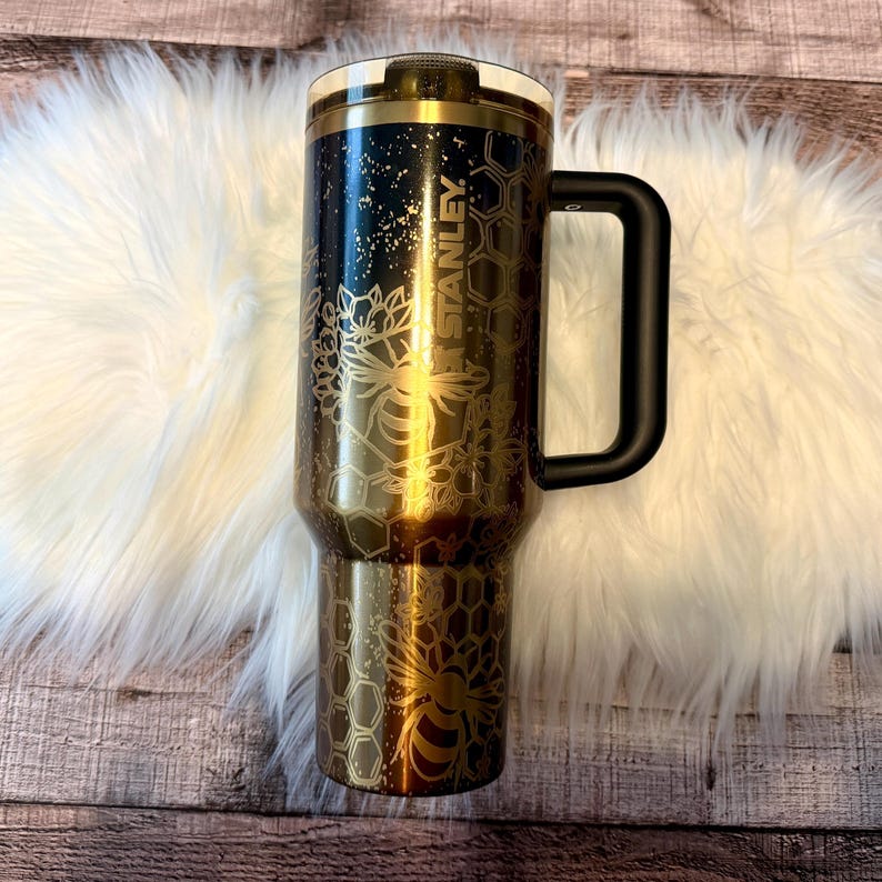 May include: A black and gold Stanley tumbler with a black handle. The tumbler features a bee and honeycomb design. The word "STANLEY" is printed vertically on the side of the tumbler. The tumbler is sitting on a white fluffy surface.