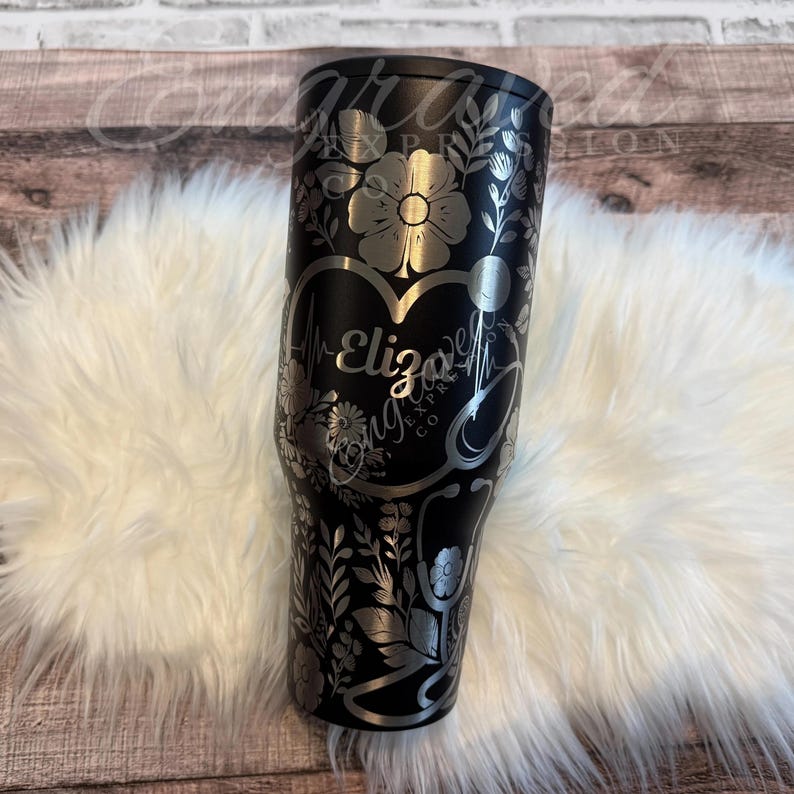 A black tumbler with a floral and heart-shaped design on it, placed on a white furry surface.