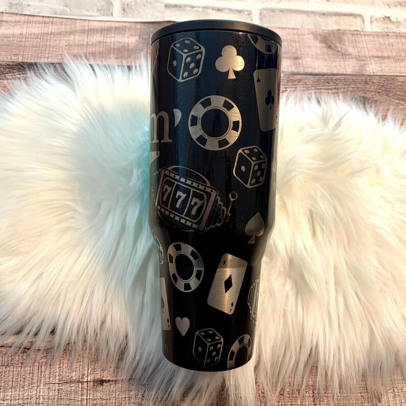 May include: Black tumbler with a black lid, decorated with silver casino-themed motifs. The design includes dice, playing cards, poker chips, and a slot machine with the number 777. The tumbler is cylindrical and appears to be made of stainless steel.