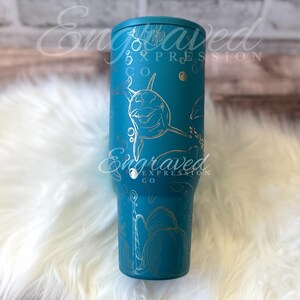 Puede incluir: Un vaso de color turquesa con un dise&ntilde;o plateado grabado que presenta delfines, burbujas y plantas acu&aacute;ticas. El vaso tiene una tapa turquesa a juego y se ven las palabras "Engraved Expression Co".