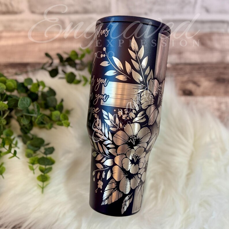 A black tumbler with a floral design on it, placed on a white fur surface next to some greenery.