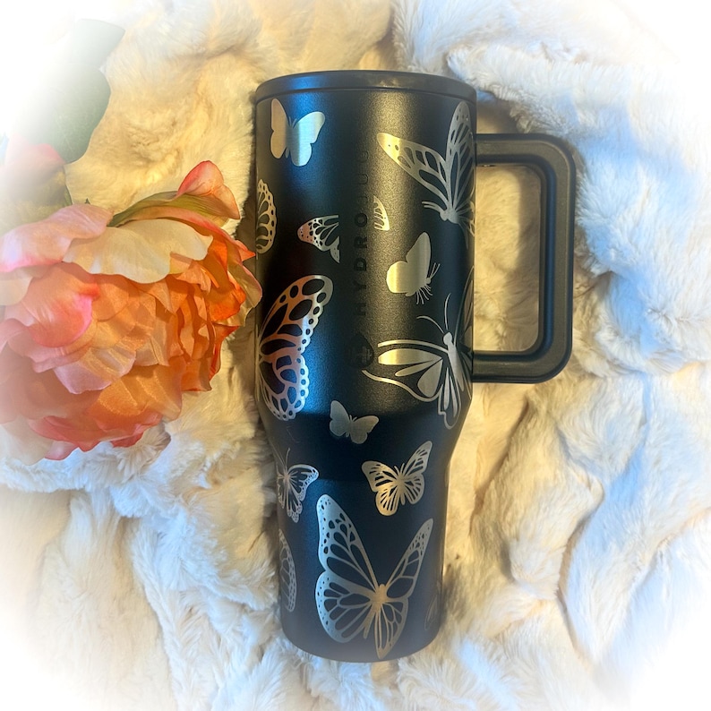 May include: A black Hydro Flask tumbler featuring a handle and silver butterfly designs. The tumbler has the Hydro Flask logo on the side and is set against a fluffy white background with a peach-colored flower.