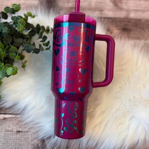 May include: A magenta insulated tumbler with a handle, lid, and straw. The tumbler is decorated with teal hearts and the word "Love" in a script font. The tumbler is sitting on a white, fluffy surface.