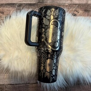 A black and silver floral patterned thermos mug with a black handle is placed on a white furry surface.