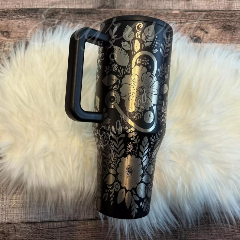 A black and silver floral patterned thermos mug with a black handle is placed on a white furry surface.