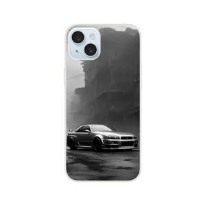 Op de afbeelding: Een zwart-witfoto van een zilveren sportwagen die door een straat in een stad rijdt. De auto is een Nissan Skyline GT-R R34. De straat wordt aan beide kanten begrensd door gebouwen. De foto is vanuit een lage hoek genomen, waardoor de kijker een goed zicht op de auto heeft.