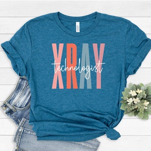 May include: Teal blue t-shirt with the word "XRAY" in colorful vertical letters and the text "technologist" in white script below.