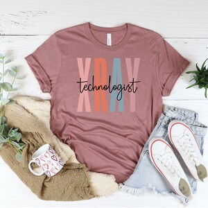 May include: A mauve t-shirt with the text "XRAY technologist" in black lettering. The letters are outlined in pink, orange, blue, and pink.