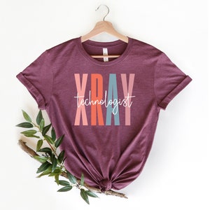 May include: A maroon t-shirt with the word "XRAY" in colorful letters and the text "technologist" in white script below.