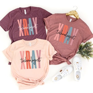 May include: Three t-shirts with the text "XRAY technologist" in colorful vertical stripes. The shirts are in different colors: maroon, pink, and light pink.