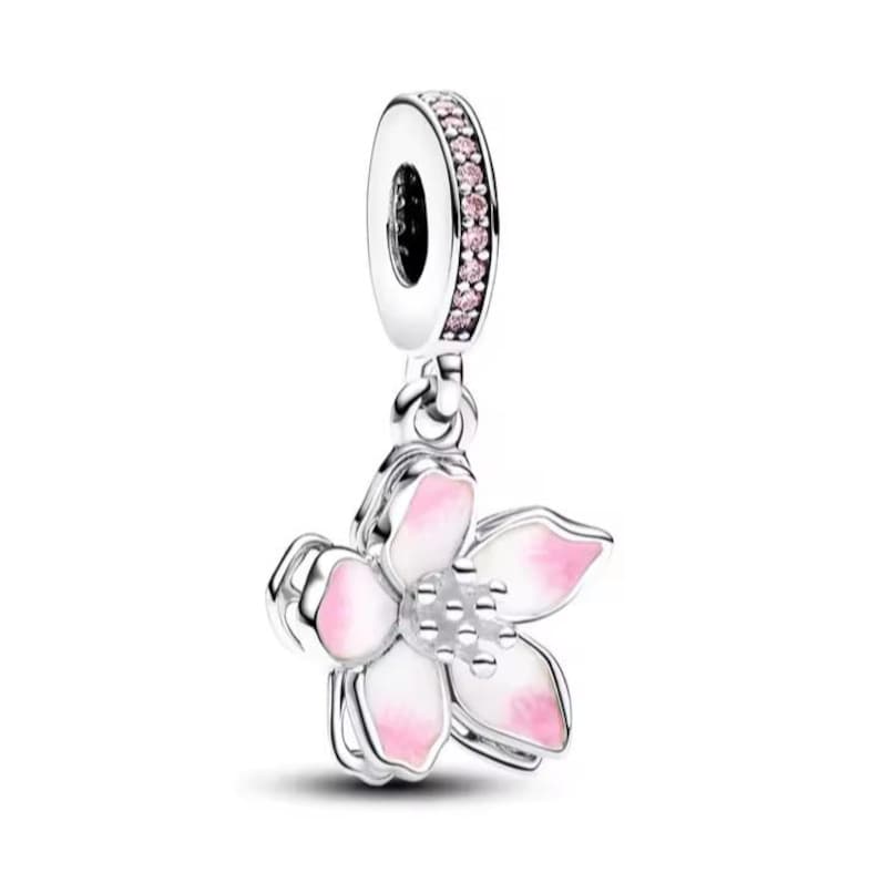 Puede incluir: Un charm de plata con un dise&ntilde;o floral. La flor tiene p&eacute;talos blancos con puntas rosas y un centro plateado. El lazo superior del charm est&aacute; adornado con piedras preciosas rosas. El charm cuelga de un lazo plateado.