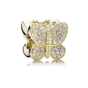 Puede incluir: Un charm de mariposa dorado con piedras transparentes y brillantes. La mariposa tiene alas detalladas y un cuerpo central, dise&ntilde;ado como accesorio de joyer&iacute;a. El charm se muestra sobre un fondo blanco.