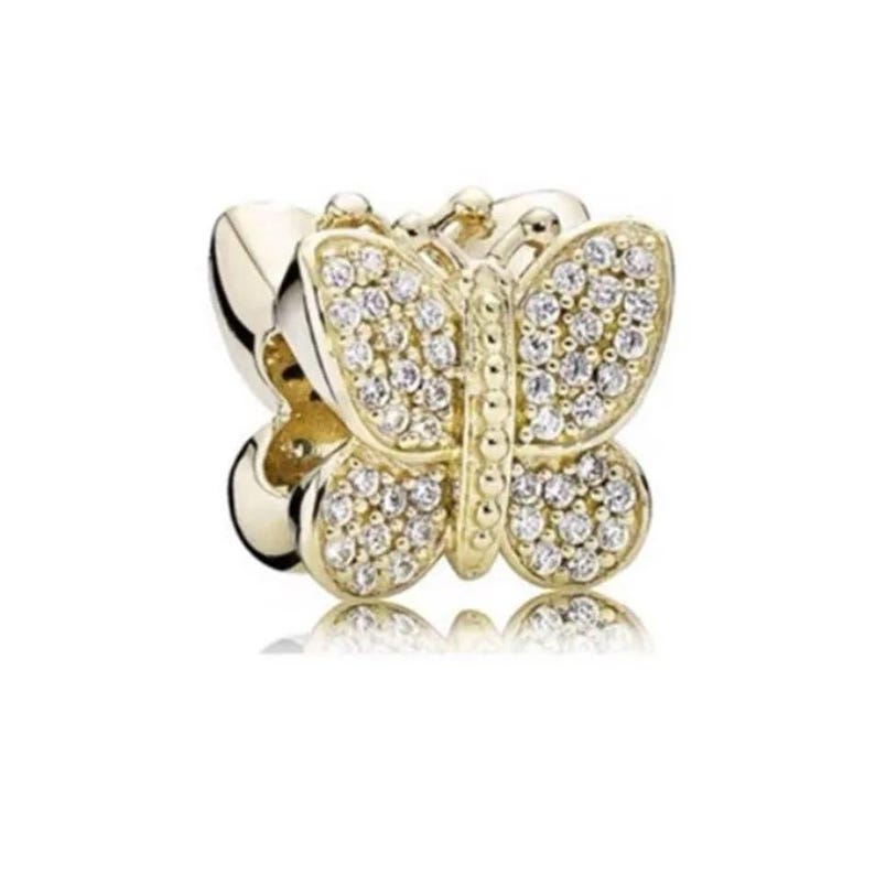 Puede incluir: Un charm de mariposa dorado con piedras transparentes y brillantes. La mariposa tiene alas detalladas y un cuerpo central, dise&ntilde;ado como accesorio de joyer&iacute;a. El charm se muestra sobre un fondo blanco.