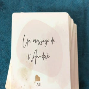 Peut inclure: Un jeu de cartes blanches aux coins arrondis, avec le texte "Un message de l'Au-delà" en écriture noire. Les cartes présentent un motif aquarelle rose clair et un logo avec le nom "Aël".