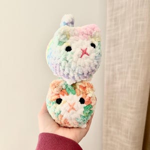 May include: Two handmade crochet cat toys. The top cat is pastel rainbow, the bottom is orange, green, and white. Both have black eyes and a pink embroidered nose. The toys are soft and fluffy.