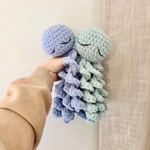 May include: Crocheted octopus toy in shades of blue and green. The toy has a round head with closed eyes and a textured body. The octopus is held against a neutral background.