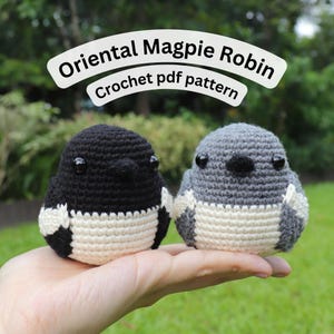 May include: Two small, round, crocheted bird figures, one black and white and one gray and white, each with black plastic eyes and a small beak. The text "Oriental Magpie Robin Crochet pdf pattern" is displayed in a white banner above the birds.