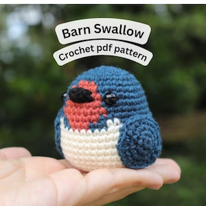 Puede incluir: Un pájaro de golondrina común de ganchillo con cuerpo azul, pecho rojo y pico negro. El pájaro tiene ojos negros y se sostiene en una mano. El texto encima del pájaro dice "Barn Swallow Crochet pdf pattern".