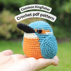 Op de afbeelding: Een gehaakte ijsvogel in blauw, oranje en zwart. De vogel heeft een zwarte snavel en een zwart oog. De tekst "Common Kingfisher Crochet pdf pattern" wordt boven de vogel weergegeven.