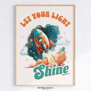 Könnte beinhalten: Gerahmter Kunstdruck mit einer Aquarellillustration einer startenden Rakete. Das Bild enthält den Text "LET YOUR LIGHT" in Orange und "Shine" in Türkis, darunter "Matthew 5:16". Die Rakete ist rot und blau.