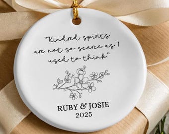 May include: A white ceramic ornament with a gold string for hanging. The ornament features the quote "Kindred spirits are not so scarce as I used to think." along with a floral design, the names "RUBY & JOSIE" and the year 2025.