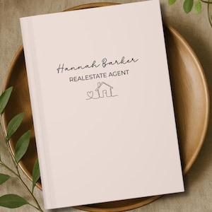 May include: A light pink notebook with the text "Hannah Barker, Real Estate Agent" and a house illustration. The notebook is on a wooden plate, with greenery and a vase of flowers in the background.