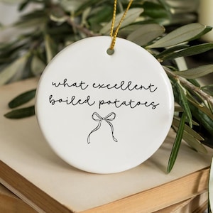 May include: A white, round ceramic ornament with the phrase "what excellent boiled potatoes" in black script, and a bow illustration. The ornament hangs from a gold string, resting on a stack of books and greenery.