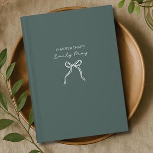 May include: A teal-colored journal with the text "CHAPTER THIRTY Emily May" and a white bow illustration. The journal is resting on a wooden plate, with greenery in the background.