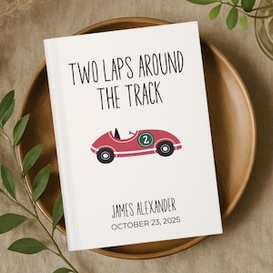 May include: A white card with the text "TWO LAPS AROUND THE TRACK" and a red race car illustration. Below the car is the name "JAMES ALEXANDER" and the date "OCTOBER 23, 2025". The card is on a wooden plate.