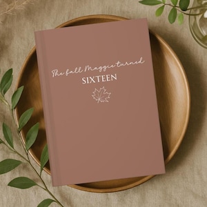 May include: A dusty rose-colored journal with the text "The fall Maggie turned SIXTEEN" in white script. A white maple leaf graphic is below the text. The journal is on a wooden plate.