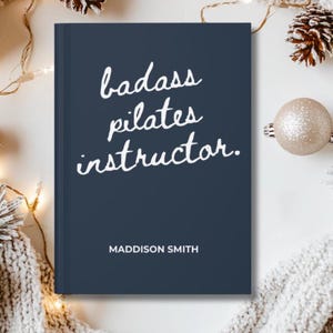 May include: A navy blue journal with the words "badass pilates instructor" in white script. The name "Maddison Smith" is printed below the text. The journal is surrounded by festive decorations.