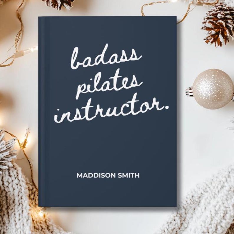 May include: A navy blue journal with the words "badass pilates instructor" in white script. The name "Maddison Smith" is printed below the text. The journal is surrounded by festive decorations.
