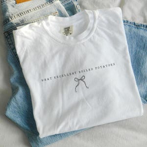 Puede incluir: Una camiseta blanca con el texto "WHAT EXCELLENT BOILED POTATOES" y un lazo dibujado. La camiseta está sobre unos vaqueros azules claros. La camiseta tiene una etiqueta Comfort Colors.